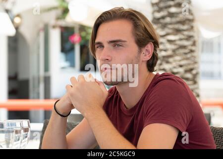 Close up of a focused handsome young man looking at the distance while sitting at an outdoor bar terrace on an out of focus background. Holidays an le Stock Photo