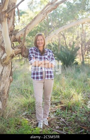 Portrait attractive mature woman in rural country, wearing plaid shirt, posing relaxed with happy smile against Australian outback bush background. Stock Photo