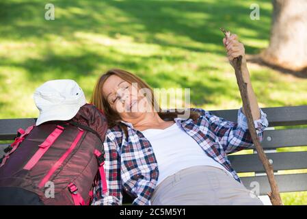 Portrait active attractive mature woman having a rest on bench, exhausted and tired after hiking, walking with backpack outdoors. Stock Photo