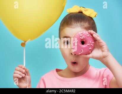 Funny little girl on background of bright blue wall. Beautiful child is having fun with balloon and donut. Yellow, pink and turquoise colors. Stock Photo