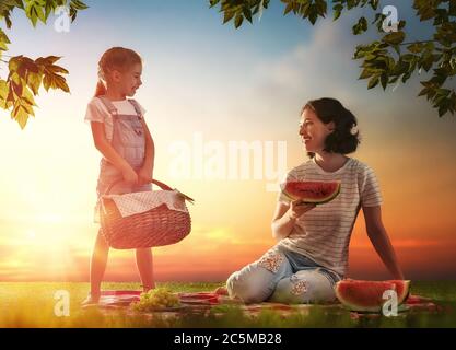 Family picnicking together. Young mother and her child daughter girl enjoying a healthy outdoor meal sitting together on green grass in summer park. Stock Photo