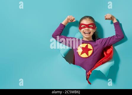 Little child is playing superhero. Kid on the background of bright blue wall. Girl power concept. Stock Photo