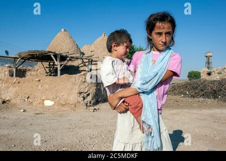 A local girl stands holding her brother in front of a beehive home in the ancient town of Harran which dates from 800 BC in eastern Turkey. Stock Photo