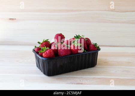 Strawberries in a dark plastic container against wood background. Sweet and beautiful berry in a plastic container. Strawberries for sale. Stock Photo