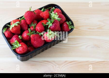 Strawberries in a dark plastic container against wood background. Sweet and beautiful berry in a plastic container. Strawberries for sale. Stock Photo
