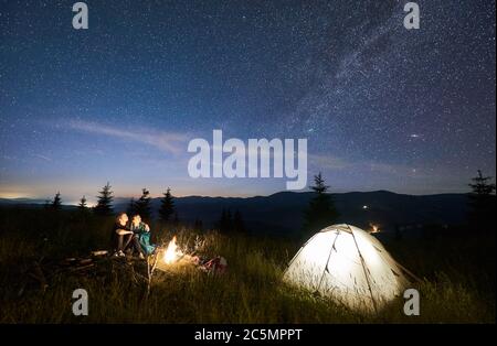 Friends hikers sitting on bench made of logs, watching amazing starry sky together in mountains beside illuminated tent, mountain ridge on background. Concept of hiking, night camping, relationships Stock Photo