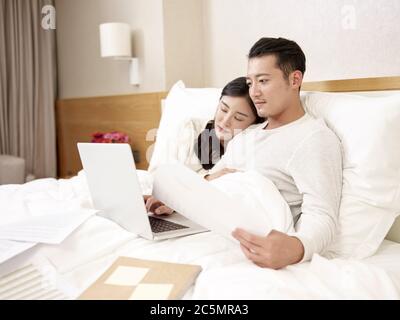 young asian business man sitting in bed working using laptop computer while wife falling asleep by his side Stock Photo