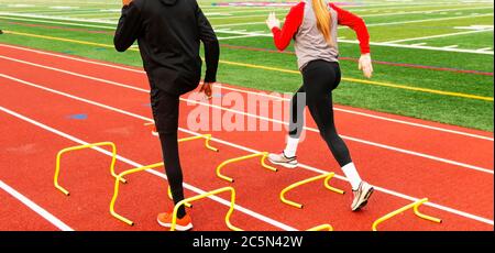 Rear view of two high school track and field athlets running over yellow mini banana hurdles in lane on a track. Stock Photo