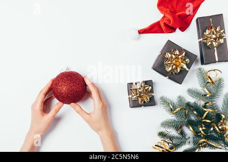 Woman's hands holding red ball Christmas toy next to fir tree branches, gift boxes, golden ribbons and Santa Claus hat on white table, flat lay. Stock Photo
