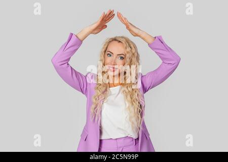 Life insurance policy. Happy adult woman with curly hair in a business suit making a gesture of a house roof over her head with her hands, feeling safe and protected, dreaming of buying a house. Stock Photo