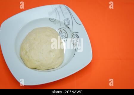 Raw dough lying on the plate on a white background. A piece of dough for bread baking . The dough is on a white plate on an orange background . Stock Photo