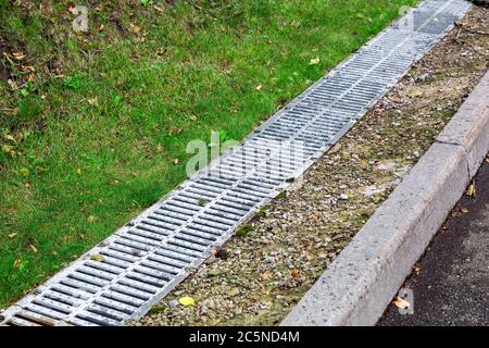 drainage system on the side of the road behind a gutter with an iron grate and green grass. Stock Photo