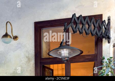 friction pantograph pendant electric heater mounted on the light wall of the street cafe with a brown window, black heater in the loft style close up. Stock Photo