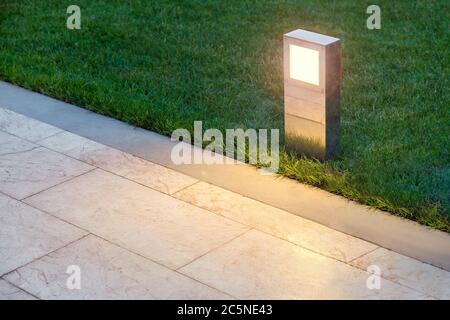 ground lamp lighting marble walkway in the evening park with a green lawn, closeup lantern illuminated marble pavement, nobody. Stock Photo