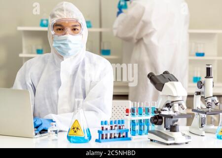 Scientists working in lab using laptop to research and develop vaccine for coronavirus covid-19 pandemic with his colleague in background. Medical Sci Stock Photo