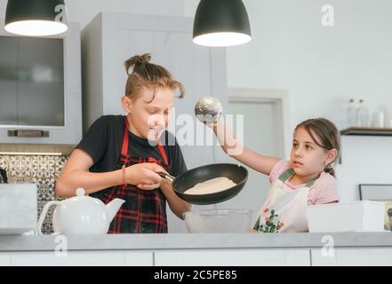 Sister and Brother dressed aprons making a homemade pancakes on the home kitchen. Girl poring a liquid dough on the hot pan. Kids Home cooking concept Stock Photo