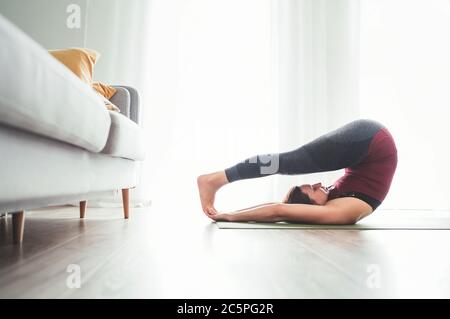 Young Woman enjoying morning yoga exercises doing Halasana pose at home living room near the big window. Active people and healthy lifestyle concept. Stock Photo