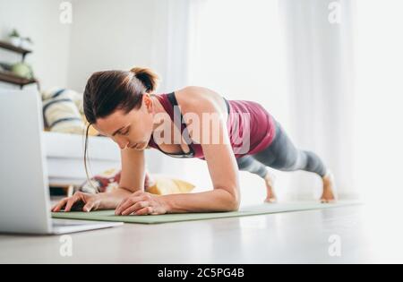 Fit sporty healthy woman on mat in low plank Chaturanga Dandasana yoga pose, doing breathing exercises, watching online yoga class on laptop computer. Stock Photo