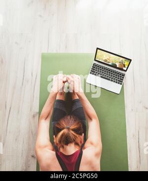 Top view at fit sporty healthy woman sitting on mat in Paschimottanasana pose, doing breathing exercises, watching online yoga class on laptop compute Stock Photo