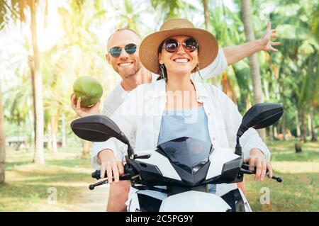 Happy smiling couple travelers riding motorbike scooter under palm trees. Laughing man carrying a coconut in hand.Tropical vacation concept image Stock Photo