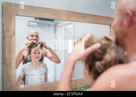 Bald dad and his long-haired teenager son in bathroom in front of the mirror. Father rising up kid's hairs thinking about his new style haircut. Funny Stock Photo