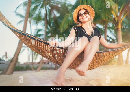 Happy smiling young woman in straw hat in sunglasses sitting in hammock and enjoy morning sun light on the Koh Samui tropical beach, Thailand. Stock Photo