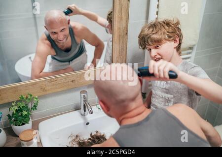 Teenager son helping his father to trim a bald head gently using electric rechargeable Trimmer in bathroom. Funny home scene and family relatives conc Stock Photo