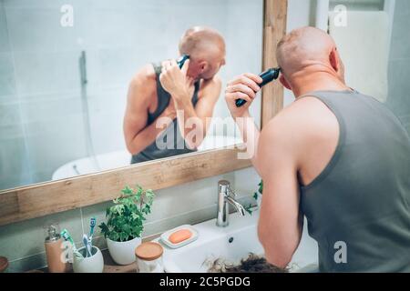 Hairless man ending shaving off all his hairs looking in bathroom mirror using an electric rechargeable Trimmer. Radical decision, image changing or b Stock Photo