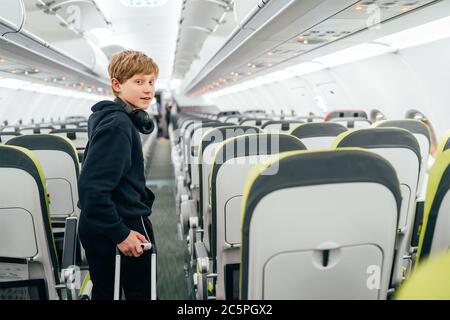 Smiling blonde hair teenager portrait staying in aircraft corridor with headphones with cabin trolley bag. Kids traveling or unaccompanied child in ai Stock Photo