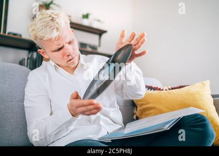 Portrait of young man sitting in comfortable sofa in living room checking out 12-inch vinyl LP record. Old vintage analog sound value concept image. Stock Photo