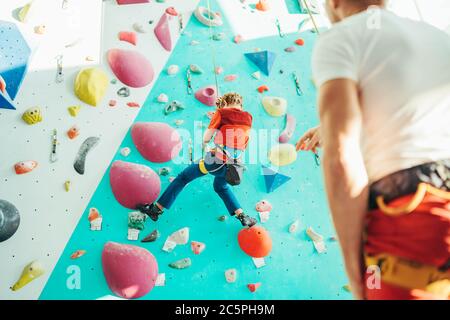 Father and teenager son at indoor climbing wall hall. Boy is hanging on the rope using a climbing harness and daddy belaying him on the floor using a Stock Photo