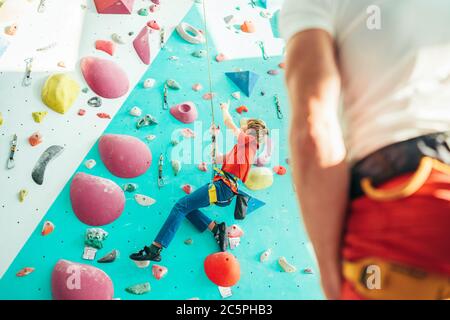 Father and teenager son at indoor climbing wall hall. Boy is hanging on the rope using a climbing harness and daddy belaying him on the floor using a Stock Photo