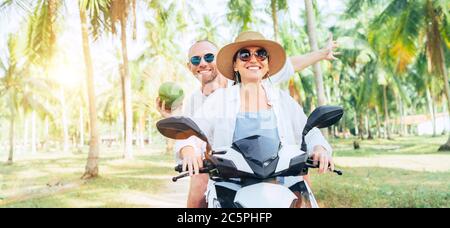 Happy smiling couple travelers riding motorbike scooter under palm trees. Laughing man carrying a coconut in hand.Tropical vacation concept image Stock Photo