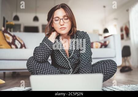 Beautiful female writer dressed pajamas typing notebook keyboard writing novel sitting cross-legged on living room floor with laptop. Distance or free Stock Photo