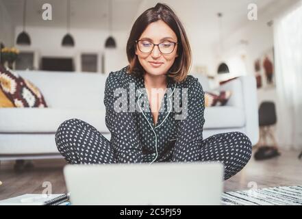 Smiling  Businesswoman dressed pajamas typing notebook keyboard chatting with colleagues sitting cross-legged on living room floor office with laptop. Stock Photo