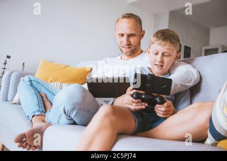 Father and son game players siting together at home on cozy sofa, using the tablet and gamepad on Coronavirus COVID-19 pandemic lockdown days. World 2 Stock Photo
