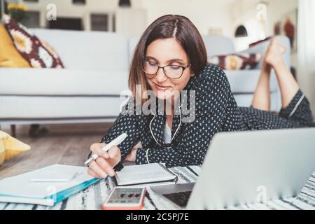 Smiling Businesswoman dressed pajamas browsing smartphone lying on living room floor "office" with laptop, papers and other stuff. Distance work in wo Stock Photo