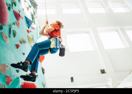 Teenager boy at indoor climbing wall hall. Boy is climbing using a top rope,chalk bag and climbing harness. Active teenager time spending concept imag Stock Photo