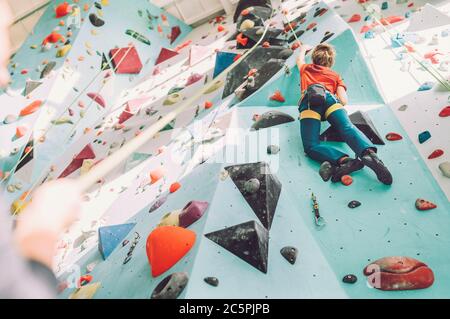 Teenager boy at indoor climbing wall hall. Boy is climbing using a top rope and climbing harness and somebody belaying him from floor. Active teenager Stock Photo