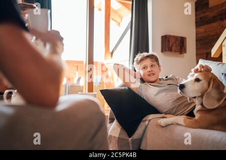 Cozy family tea time. Father and son at the home living room. Boy lying on comfortable sofa and  stroking their beagle dog and smiling. Peaceful famil Stock Photo