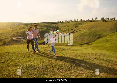 Adorable little girl flying kite while her parents walking behind her in mountains during summer camping trip, copy space. Family vacation Stock Photo