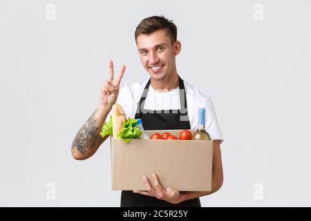 Retail, grocery shopping and delivery concept. Charismatic handsome salesman, courier in black apron show peace sign and holding box with groceries Stock Photo