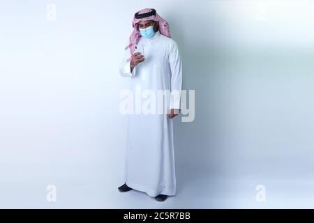 Saudi Arab Man standing and using mobile phone while wearing face mask Stock Photo