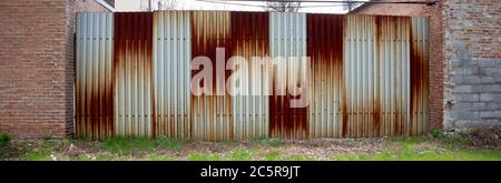 Rusted corrugated sheet metal fence between two brick walled buildings. Stock Photo
