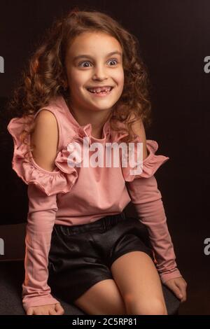 Little girl in pink blouse and tight shorts on dark background smiles, shows no tooth Stock Photo