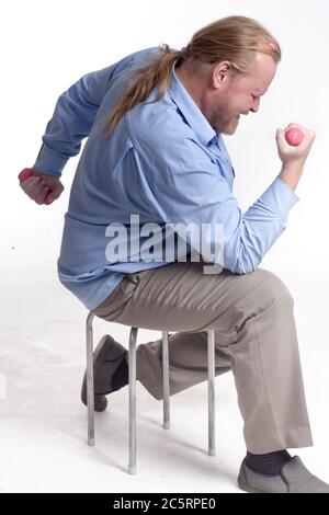 a stout, middle-aged, unsportsmanlike man in a blue shirt with long hair sits on a chair and lifts pink, light-weight dumbbells Stock Photo