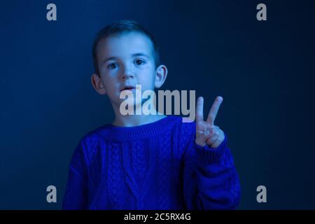 Studio portrait of a brunette Boy who counts on his fingers, shows two finger, toning in a classic blue color. Stock Photo
