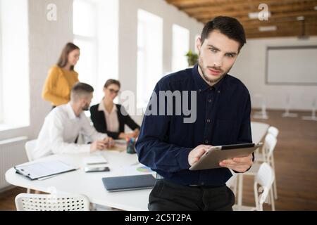 Young business man with beard in shirt thoughtfully looking in camera while holding tablet in hands in office with colleagues on background Stock Photo