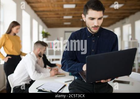 Young man with beard in shirt thoughtfully using laptop while spending time in office with colleagues on background Stock Photo