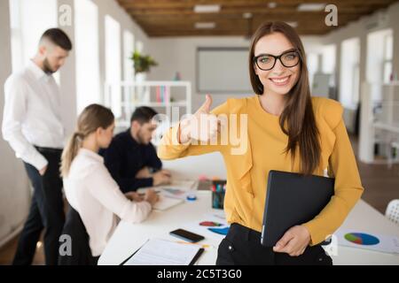 Young smiling business woman in eyeglasses happily looking in camera while showing thumb up gesture with folder in hand in office with colleagues on Stock Photo